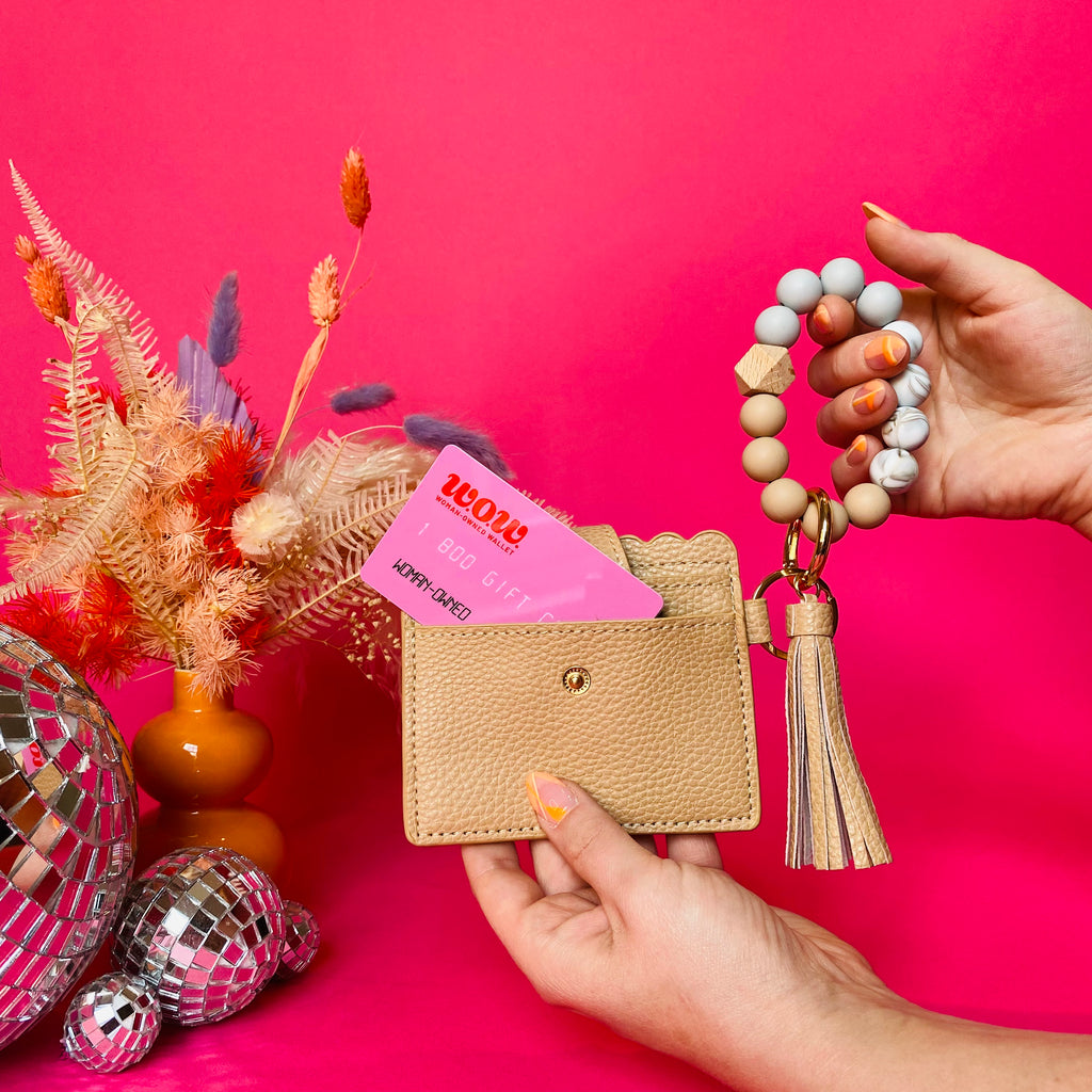 Closeup of woman's hands holding a tan faux leather wallet featuring two back card pockets, a snap closure, attached bracelet made from grey and tan silicone beads, and a tan faux leather tassel. There is a pink gift card from Woman-Owned Wallet in the ID pocket. Pink backdrop with an orange vase filled with dried florals sitting next to small disco balls.