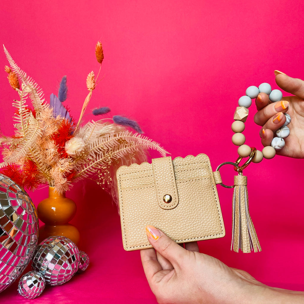 Closeup of woman's hands holding a tan faux leather wallet featuring two back card pockets, a snap closure, attached bracelet made from grey and tan silicone beads, and a tan faux leather tassel. Pink backdrop with an orange vase filled with dried florals sitting next to small disco balls.