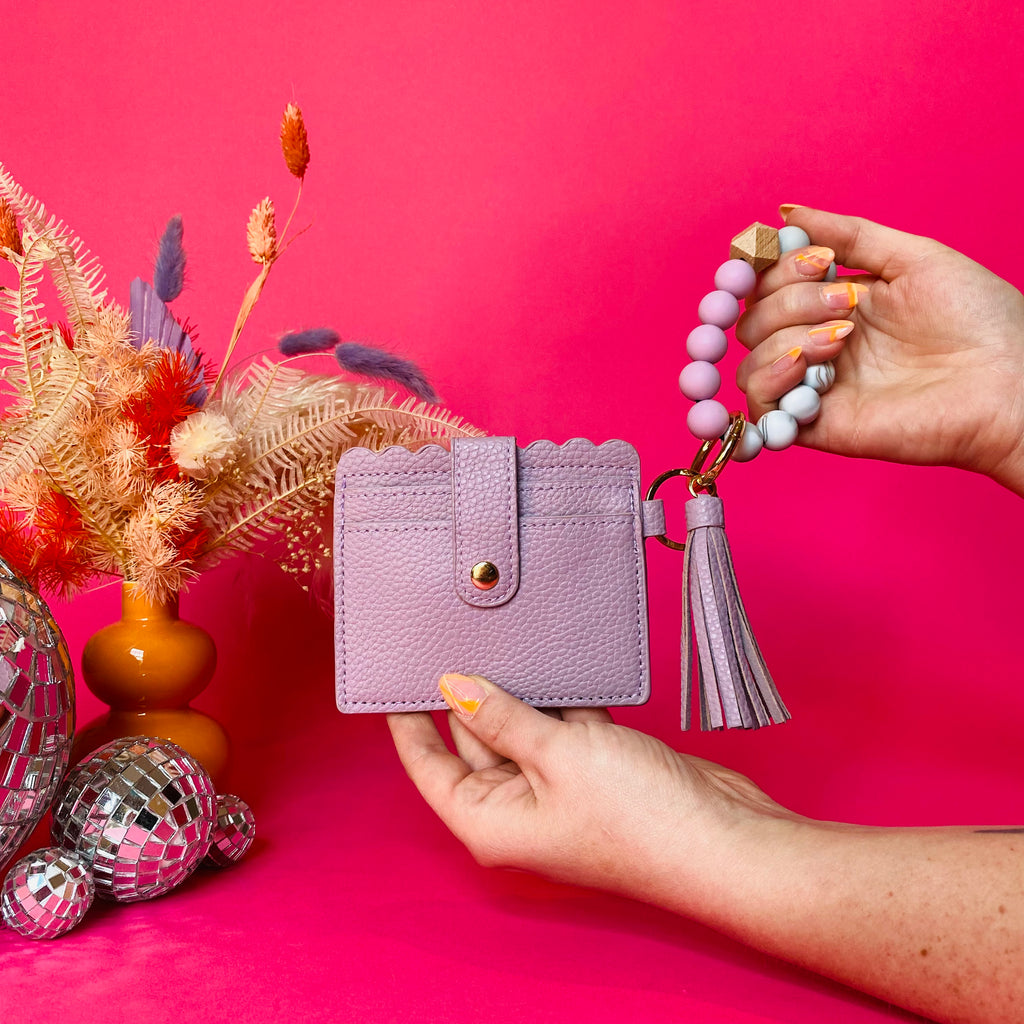 Closeup of woman's hands holding a purple faux leather wallet featuring two back card pockets, a snap closure, attached bracelet made from grey and purple silicone beads, and a purple faux leather tassel. Pink backdrop with an orange vase filled with dried florals sitting next to small disco balls.