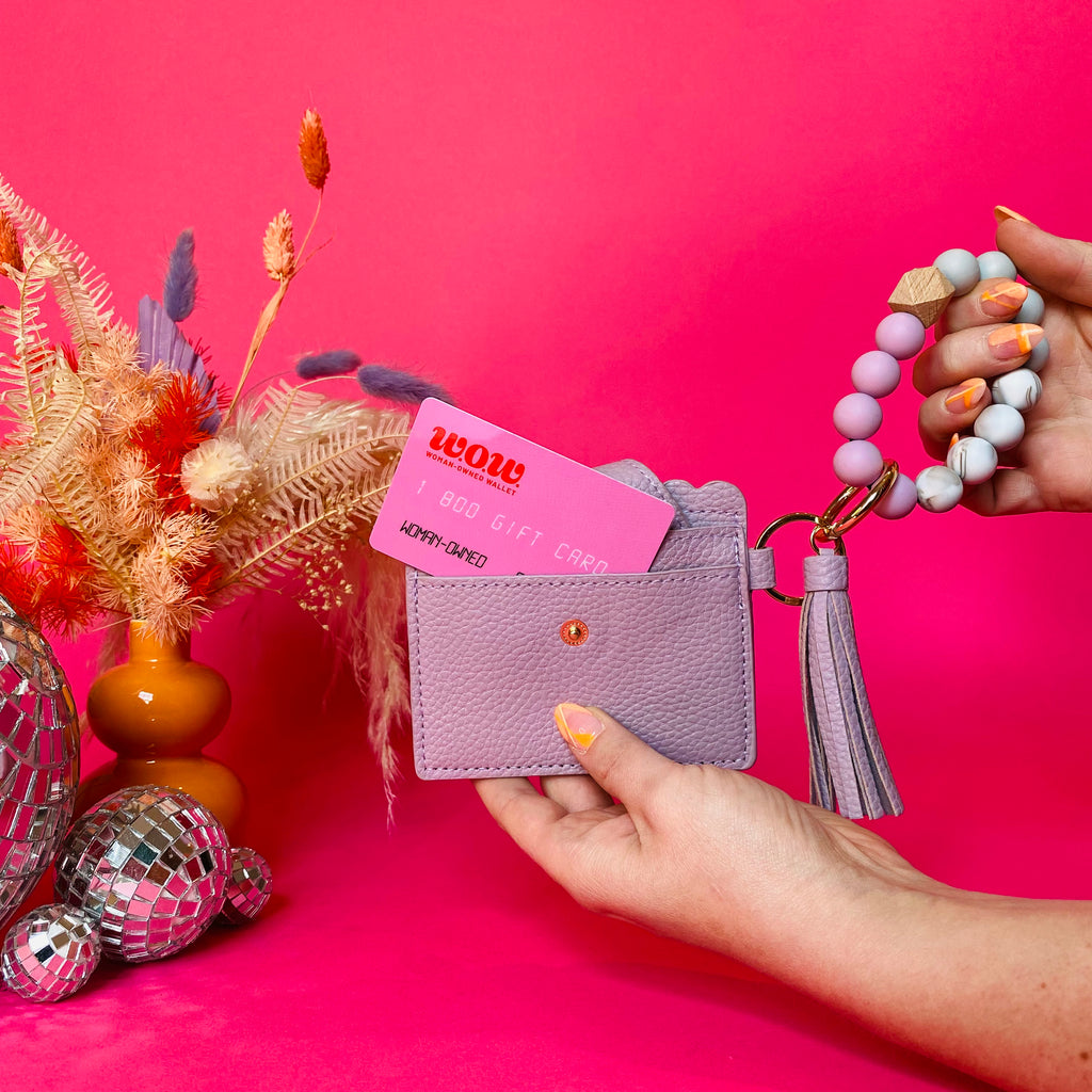 Closeup of woman's hands holding a purple faux leather wallet featuring two back card pockets, a snap closure, attached bracelet made from grey and purple silicone beads, and a purple faux leather tassel. There is a pink gift card from Woman-Owned Wallet in the ID pocket. Pink backdrop with an orange vase filled with dried florals sitting next to small disco balls.