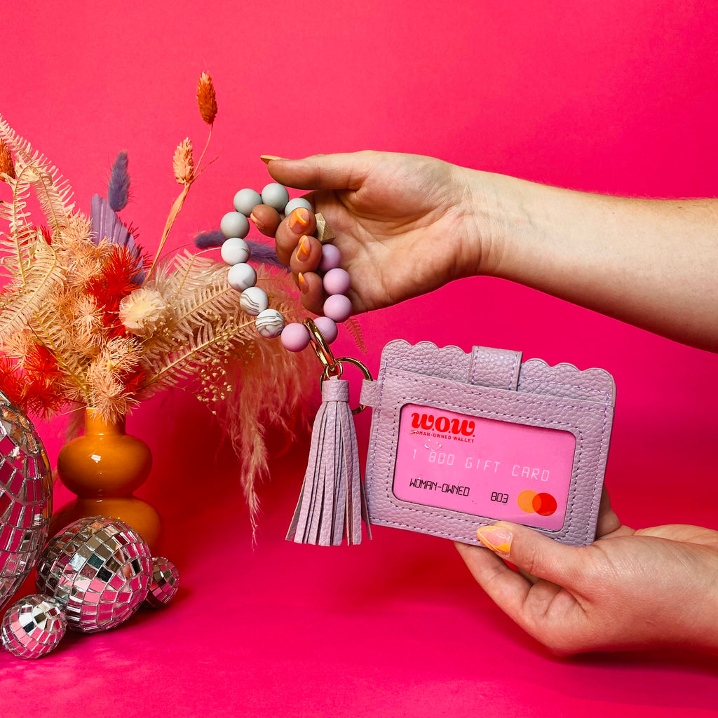 Closeup of woman's hands holding a purple faux leather wallet featuring a clear ID pocket, attached bracelet made from purple and white silicone beads, and a purple faux leather tassel. There is a pink gift card from Woman-Owned Wallet in the ID pocket. Pink backdrop with an orange vase filled with dried florals sitting next to small disco balls.