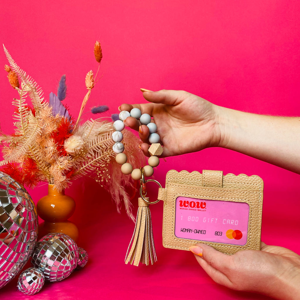 Closeup of woman's hands holding a tan faux leather wallet featuring a clear ID pocket, attached bracelet made from grey and tan silicone beads, and a tan faux leather tassel. There is a pink gift card from Woman-Owned Wallet in the ID pocket. Pink backdrop with an orange vase filled with dried florals sitting next to small disco balls.